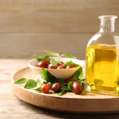Flasche mit goldenem Simmondsia Chinensis Seed Oil neben einer Schale mit Jojobasamen und grünen Blättern auf einem Holzteller.