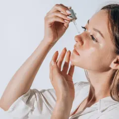 Frauenhand trägt Tropfen eines Serums auf das Gesicht auf. Ob Squalan oder Hyaluron, die Haut strahlt Feuchtigkeit aus.