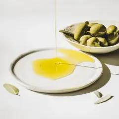 Flüssiges Olea europaea fruit oil fließt auf einen Teller, daneben eine Schale mit grünen Oliven und Blättern.
