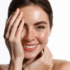 Strahlendes Lächeln einer Frau mit Sommersprossen, die sich die Hand vors Gesicht hält. Sie fragt sich, was ist kollagen und wie es ihre Haut jugendlich hält.