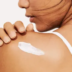 Eine Hand trägt eine weiße Creme auf die Schulter auf. Die Creme könnte Panthenol oder Bepanthen enthalten, um die Haut zu pflegen.