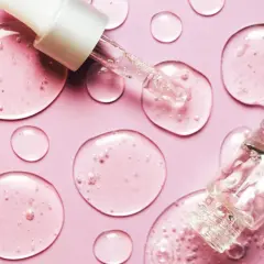 Tropfen von natürlichem Hyaluron auf rosa Untergrund. Eine Pipette und ein Fläschchen sind zu sehen.