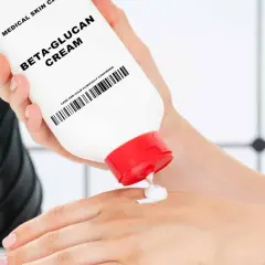Eine Hand hält eine Tube "Beta-Glucan Cream" und gibt eine kleine Menge auf die andere Hand. Die Creme verspricht eine positive **Beta-Glucan Wirkung Haut**.