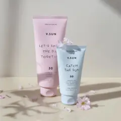 Zwei V.SUN Sonnencreme-Tuben, eine mit LSF 30 für den Körper, die andere mit LSF 50 für das Gesicht, umgeben von zarten Blütenblättern.