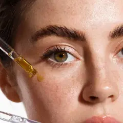 Hände tragen Pipetten mit Seren auf das Gesicht einer Frau auf. Diese Seren enthalten oft Peptide, die für die Hautpflege wichtig sind.
