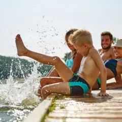 Familie genießt den Sommer am See. Kinder planschen im Wasser, Eltern entspannen sich. Guter uva schutz ist wichtig.