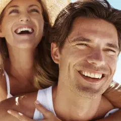 Glückliches Paar am Strand, die Frau lacht und trägt einen Hut. Selbst im Schatten wird man braun, wenn man die Sonne genießt.