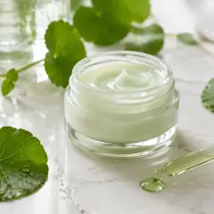 Grüner Tiegel mit Cica-Creme und Pipette, umgeben von frischen Blättern. Cica ist bekannt für seine beruhigenden Eigenschaften.