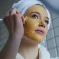 Junge Frau mit Handtuch auf dem Kopf trägt eine Kurkuma-Maske auf. Vorher-Nachher-Effekt für strahlende Haut.