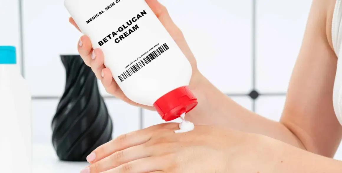 Eine Hand hält eine Tube "Beta-Glucan Cream" und gibt eine kleine Menge auf die andere Hand. Die Creme verspricht eine positive **Beta-Glucan Wirkung Haut**.